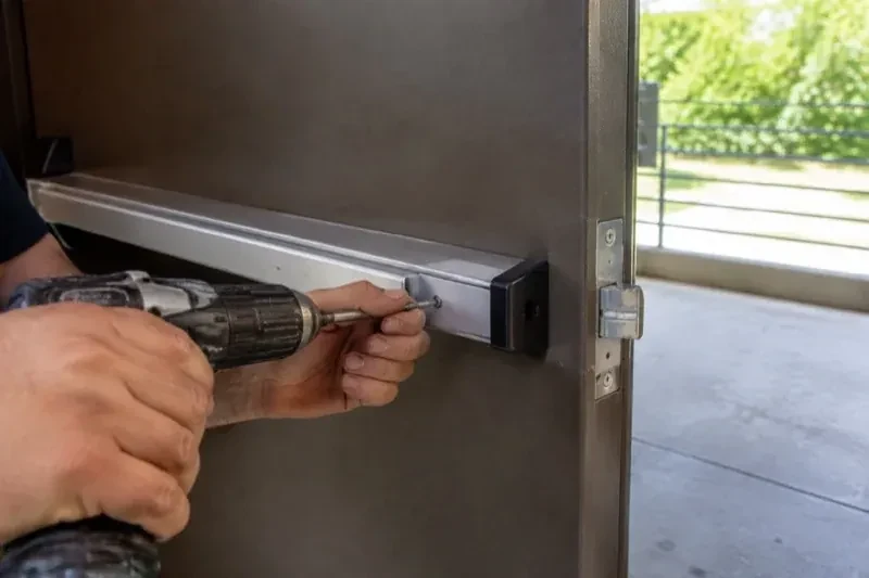 Hands using a power driver at the end of a horizontal push bar on an open metal door. — Panic & Exit Hardware, London