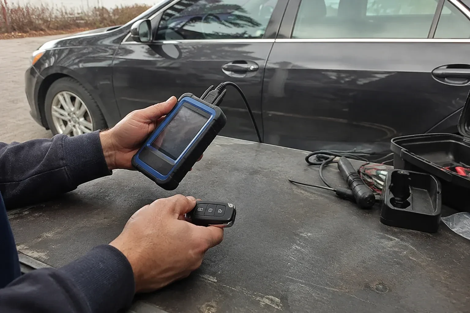 Hands holding a blue-edged electronic device and car-key fob at a worktable beside a parked car. — Transponder Key Programming, London